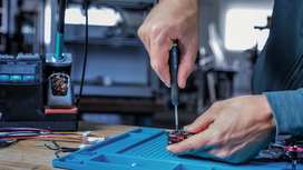 Person uses a screwdriver to assemble an electronic component on a blue mat.