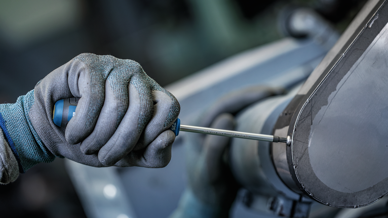 Person wearing safety equipment uses a screwdriver to tighten a machine panel.