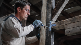 Person wearing safety equipment fastens a metal bracket to a wooden beam.