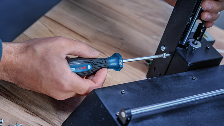 Person using a screwdriver to fasten a metal bracket on a wooden surface.