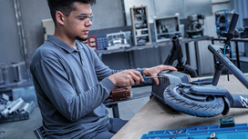 Person wearing safety equipment repairs an electric scooter at a workshop table.