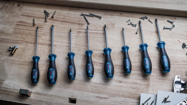 Eight screwdrivers with blue handles arranged on a wooden workbench near scattered screws.