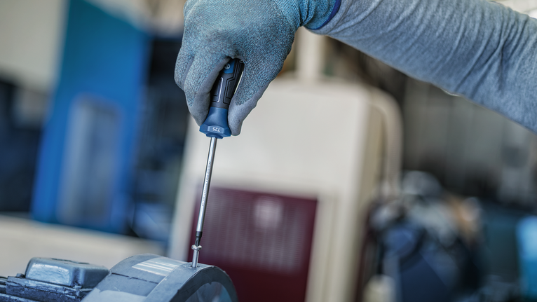 Person wearing safety equipment tightens a screw on machinery with a screwdriver.
