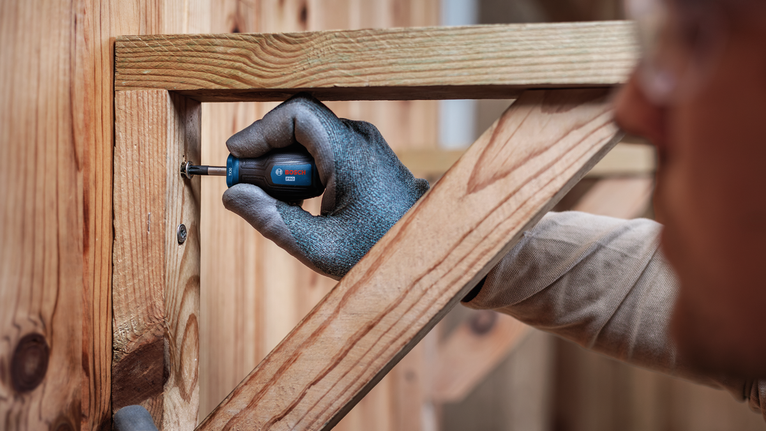 Person wearing safety equipment fastens a screw into a wooden frame.