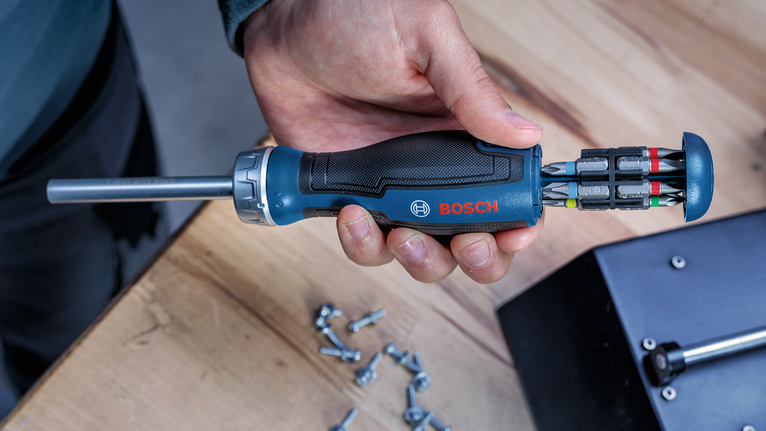 Person holding a screwdriver with multiple bits over a wooden workbench.