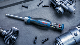 Multi-bit screwdriver with color-coded bits lies on a workbench among automotive parts and bolts.