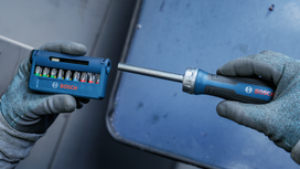 Person wearing safety equipment holds a screwdriver and bit set above a metal surface.