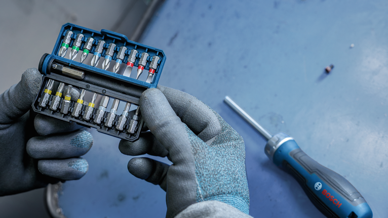 Person wearing safety equipment holding a bit set next to a screwdriver on a workbench.