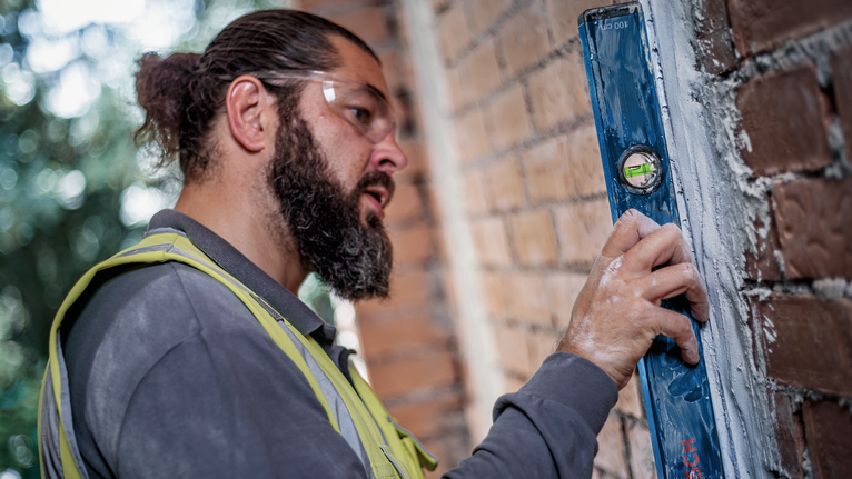 A person wearing safety equipment checks wall alignment with a blue spirit level.