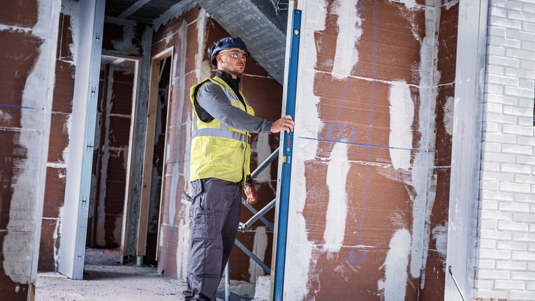 A person wearing safety equipment checks a metal frame alignment inside a building under construction.