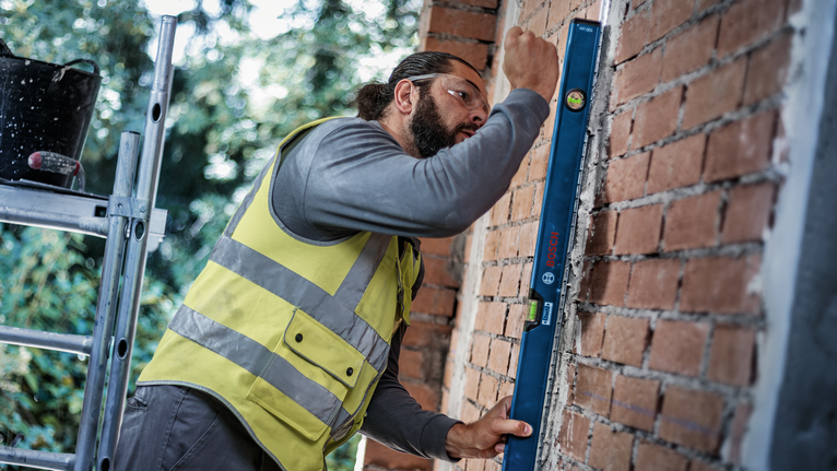 Person wearing safety equipment checks brick wall alignment with a blue spirit level.