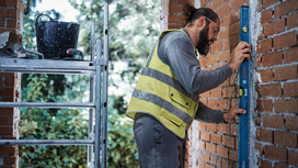 A person wearing safety equipment checks brick wall alignment with a spirit level.