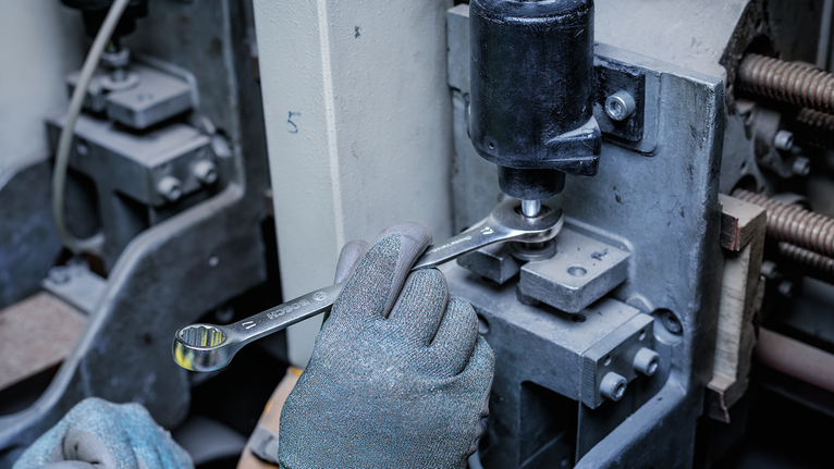 Person wearing safety equipment uses a wrench to tighten a bolt on industrial machinery.