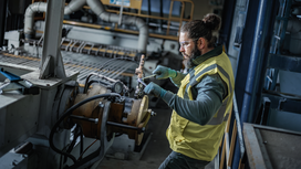 Person wearing safety equipment uses a power tool to adjust machinery in an industrial setting.