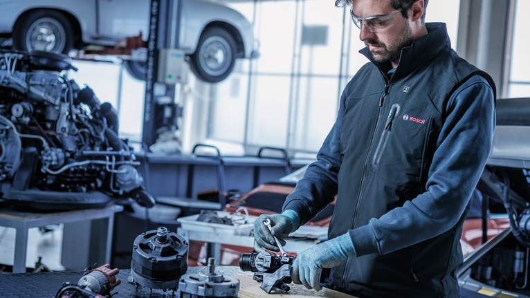 Person wearing safety equipment repairs a car alternator in an automotive workshop.