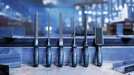 Six metal files with black and blue handles are arranged upright on a metal beam in a workshop.
