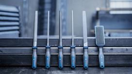 Six metal files with blue handles arranged on a metal beam in a workshop.