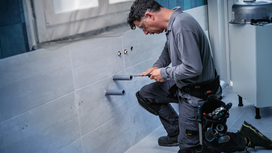 Person wearing safety equipment working on wall pipes in a tiled bathroom.