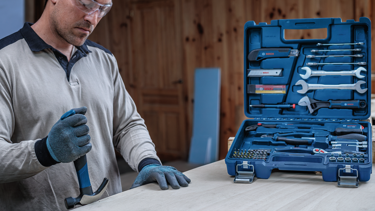 Person wearing safety equipment uses a hammer next to an open toolkit on a table.