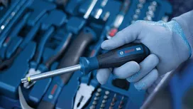 Person wearing safety equipment holds a screwdriver above a toolkit.