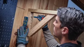 Person wearing safety equipment uses a screwdriver to attach a wooden frame to a wall.