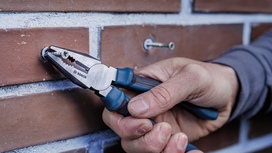 Person uses pliers to hold a screw against a brick wall.