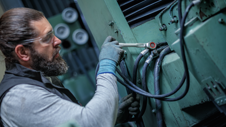 Person wearing safety equipment tightens a bolt on industrial machinery.