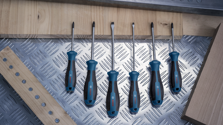 Six screwdrivers with blue and black handles arranged on a metal surface near wood pieces.