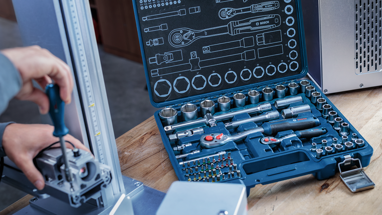 Toolkit with socket wrenches and bits open on a table, person using a screwdriver nearby.