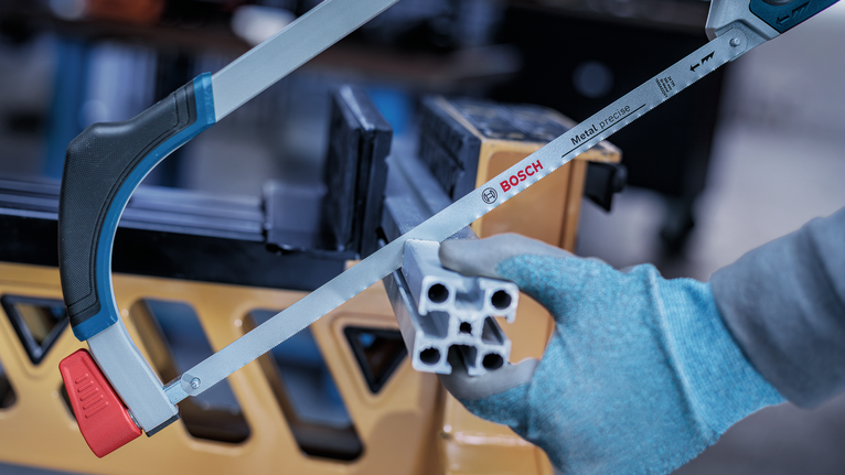 Person wearing safety equipment uses a hacksaw to cut an aluminum profile.