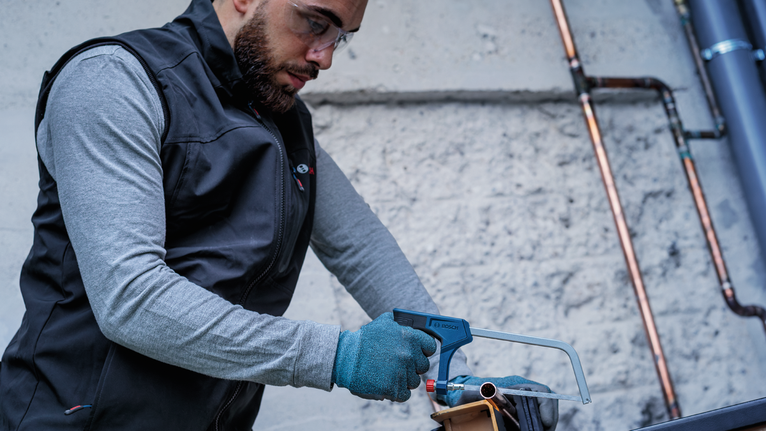 Person wearing safety equipment uses a hacksaw to cut a metal pipe.