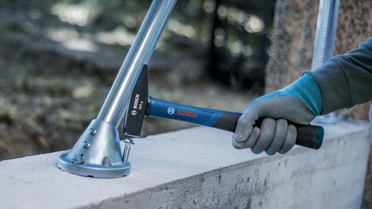 Person wearing safety equipment uses a hammer to secure a metal support on a concrete surface.