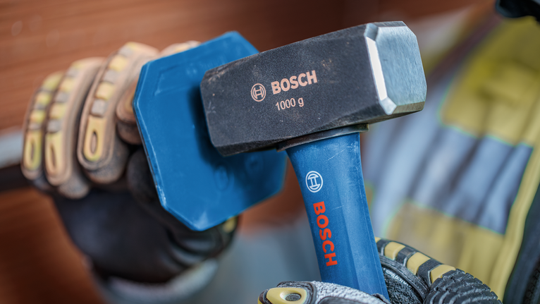 Person wearing safety equipment strikes a metal chisel with a heavy hammer.