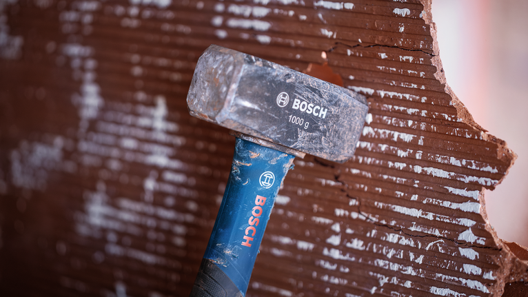 A sledgehammer breaking a grooved red brick wall.