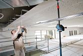 A person wearing safety equipment installs metal framing while using a laser leveling tool.