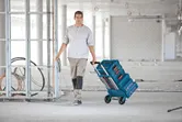 A person pulls a collapsible hand truck with stacked toolboxes in a construction area.