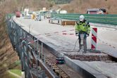 A person wearing safety equipment operates a breaker to demolish concrete on a bridge.