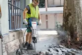 A person wearing safety equipment breaks pavement outdoors with a large electric breaker.