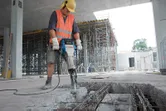 A person wearing safety equipment uses a breaker to demolish concrete at a construction site.
