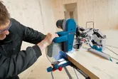 A person wearing safety equipment cuts a wood plank with a sliding mitre saw.