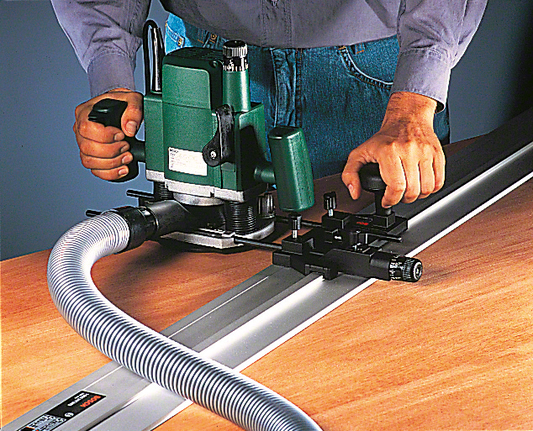 Person guides a power router along a rail to cut wood, dust hose attached.
