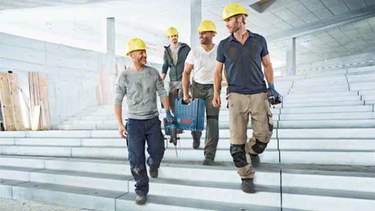 Four workers in yellow helmets walk down concrete stairs, carrying Bosch tools.